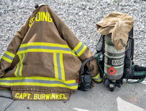 A firefighter's jacket labeled "CAPT. BURWINKEL" and reflective stripes is draped next to a helmet and an oxygen tank on a sidewalk, with gravel in the background.