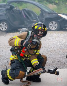A firefighter in full gear kneels on the ground holding a fire hose nozzle, with a damaged car and debris visible in the blurred background.
