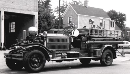 A vintage fire truck with a uniformed driver is parked outside a fire station. The truck features a ladder and various firefighting equipment, and a residential house is visible in the background.